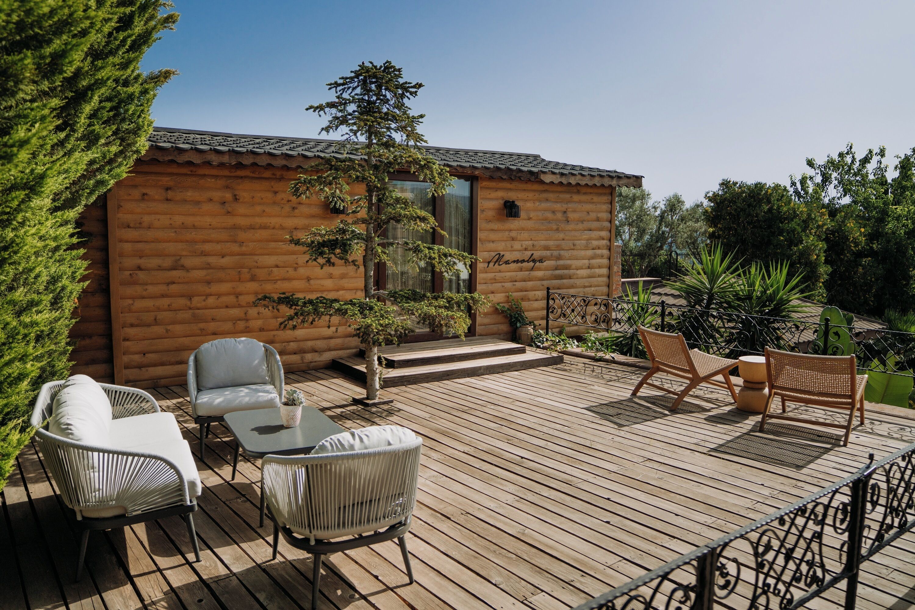 Una cabaña de madera con terraza, muebles de mimbre blancos y un pequeño jardín.