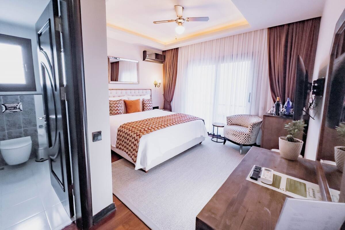 A modern hotel room with a large bed, a sitting area with a chair, a desk, and a ceiling fan.