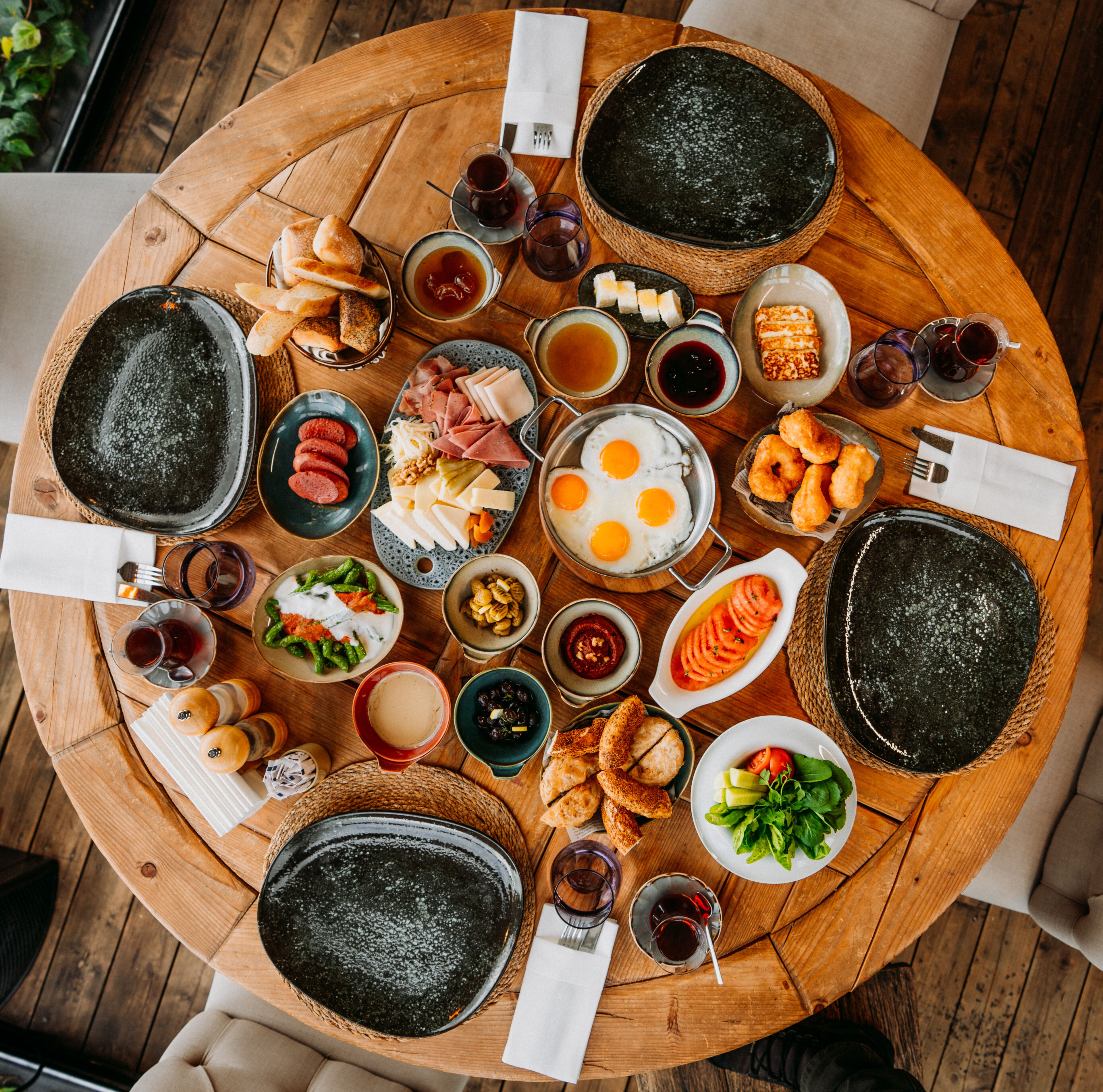 Ein runder Holztisch mit verschiedenen Speisen und Getränken, darunter Käse, Fleisch und Gemüse, für eine Mahlzeit angerichtet.