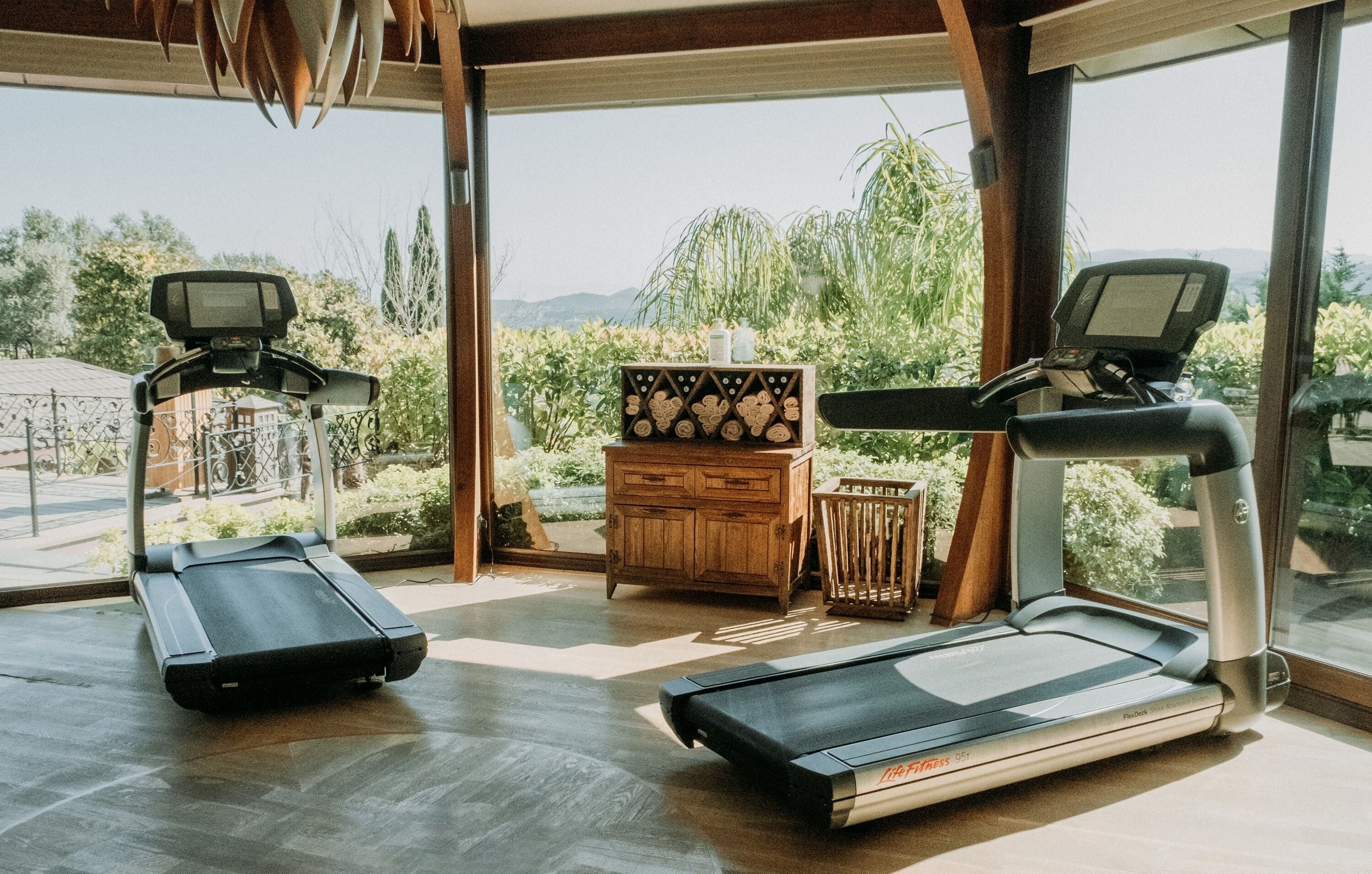A home gym with two treadmills, a wooden cabinet, and a view of greenery and mountains through large windows.