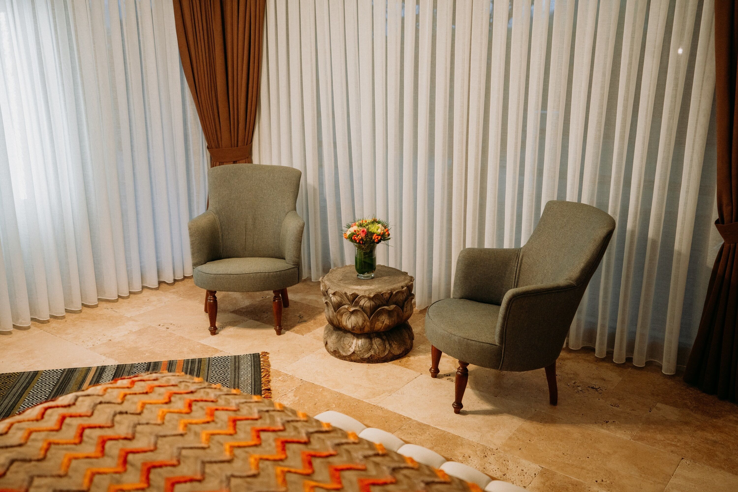 A room with two armchairs, a round wooden side table with a vase of flowers, and a patterned rug on a tiled floor.