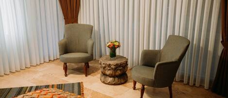 A room with two armchairs, a round wooden side table with a vase of flowers, and a patterned rug on a tiled floor.