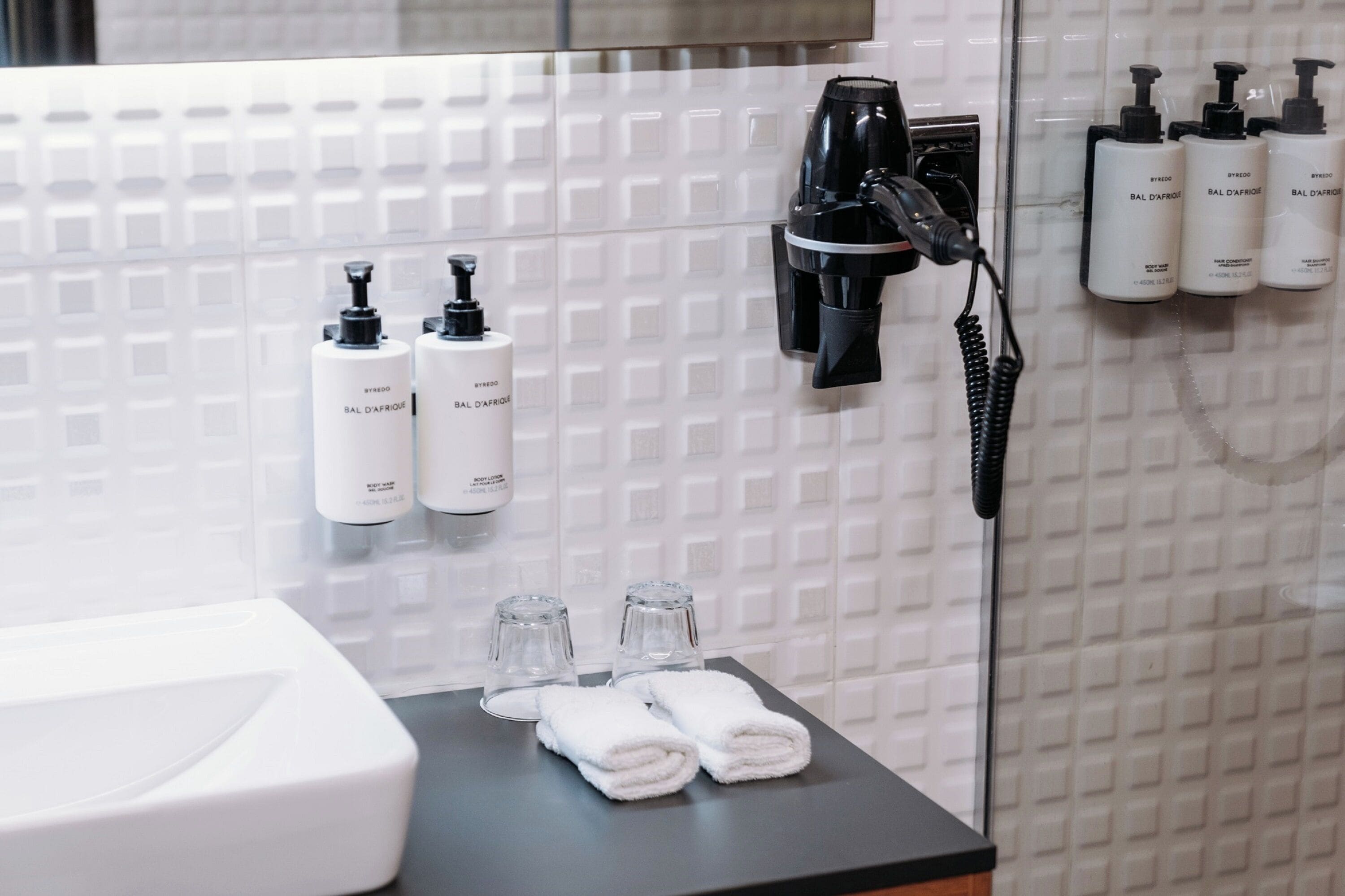 A bathroom with a white tiled wall, a hairdryer, and three bottles of Bal D'Afrique shampoo. There are two towels and two glasses on the counter.