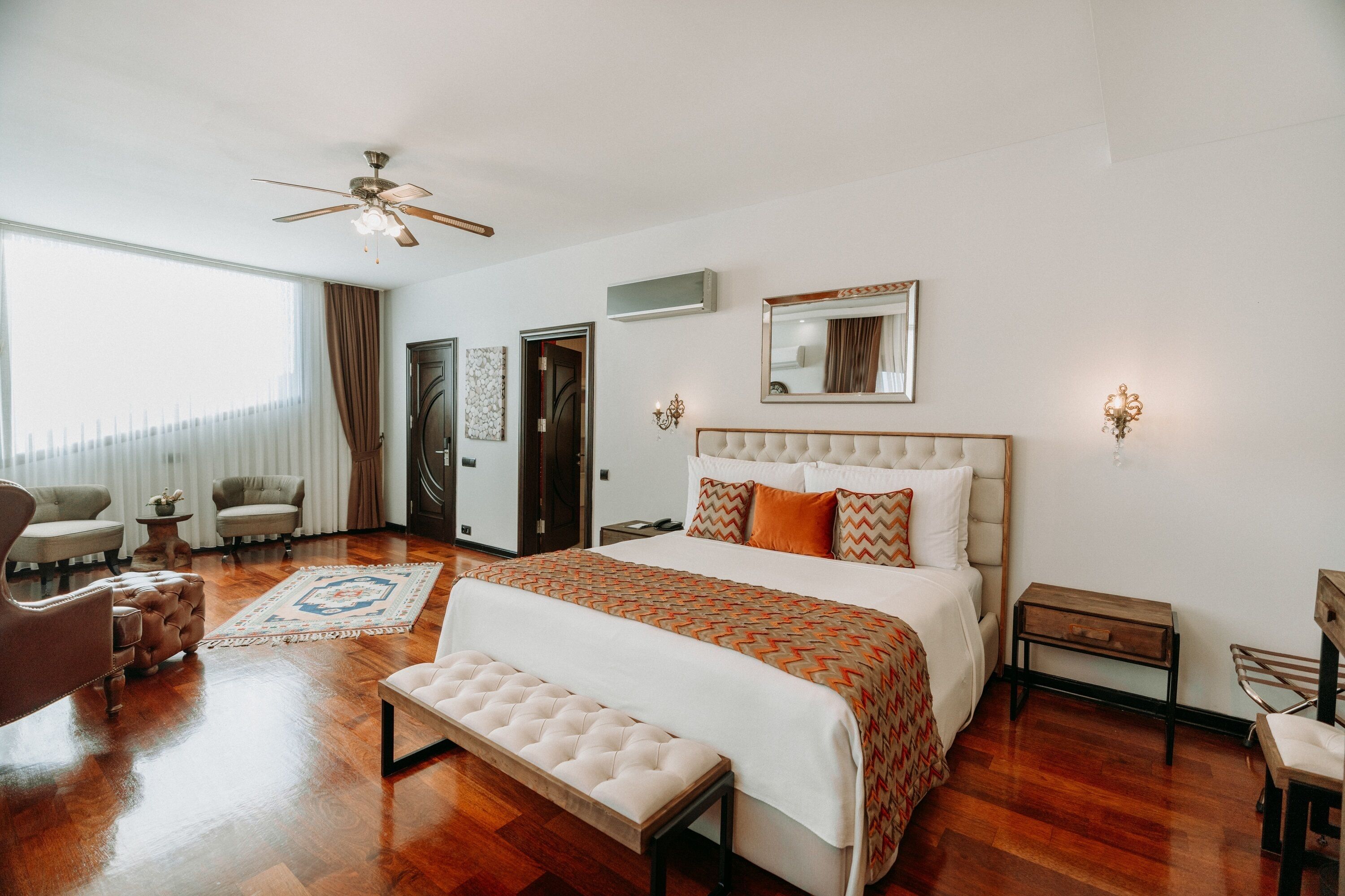 A hotel room with a large bed, a ceiling fan, a sitting area with chairs, a small table, and a bench.