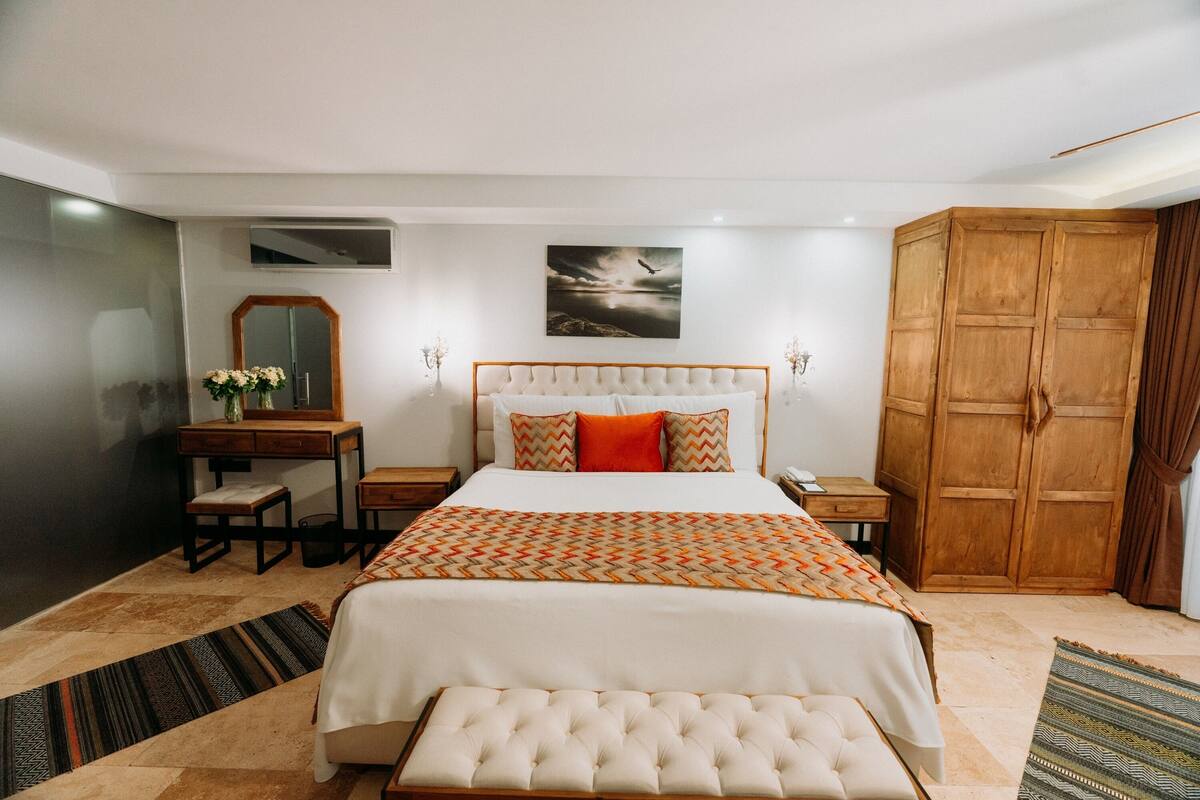 A bedroom with a large bed, a wooden wardrobe, a desk with a mirror, and a painting on the wall.
