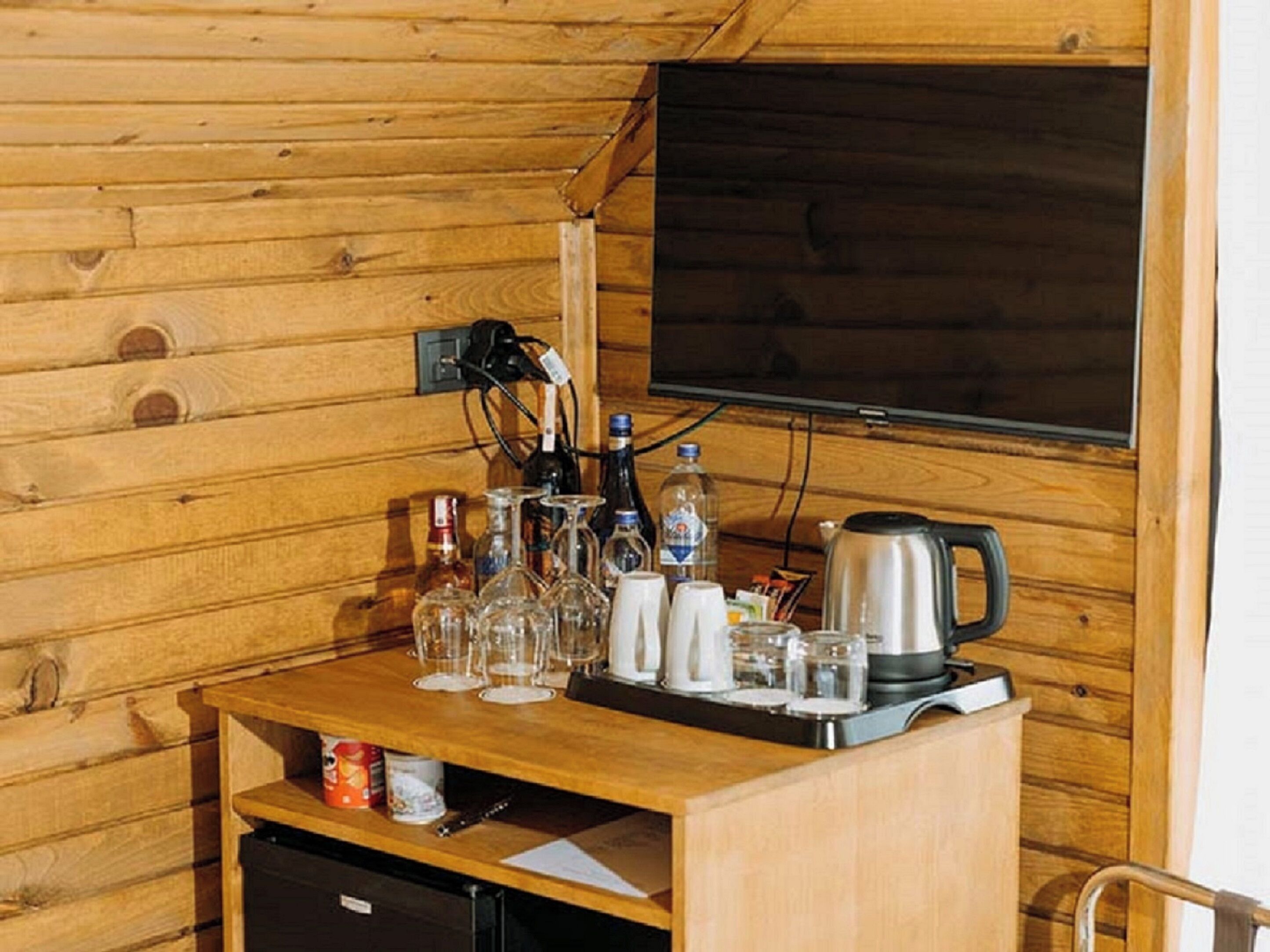 A wooden cabin interior with a wooden table set with glasses, bottles, and a kettle. A television is mounted on the wall above the table.