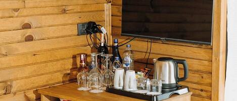 A wooden cabin interior with a wooden table set with glasses, bottles, and a kettle. A television is mounted on the wall above the table.