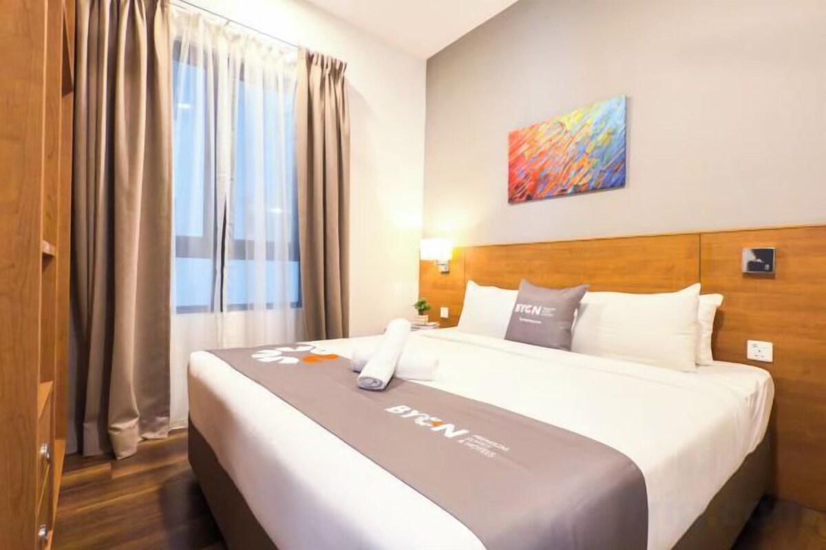 Una habitación de hotel con una cama grande, cabecero, ventana con cortinas, una obra de arte en la pared y una mesita de noche con lámpara.