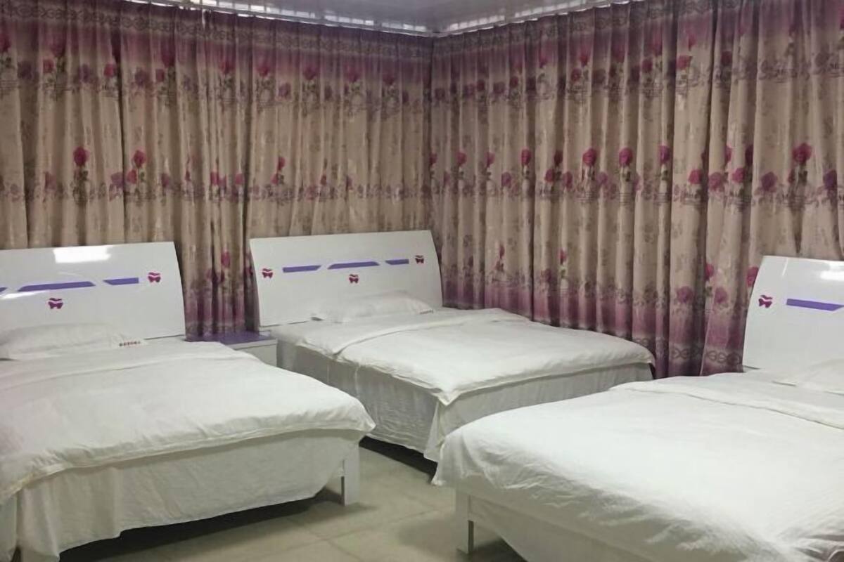A hotel room with three single beds, a large window with curtains, and a plain wall.