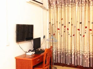 A room with a flat-screen TV mounted on the wall, a wooden desk with a computer, and a curtain with a floral pattern. - Hezhou Huangyao Longquan Business Hotel (Zhaoping)