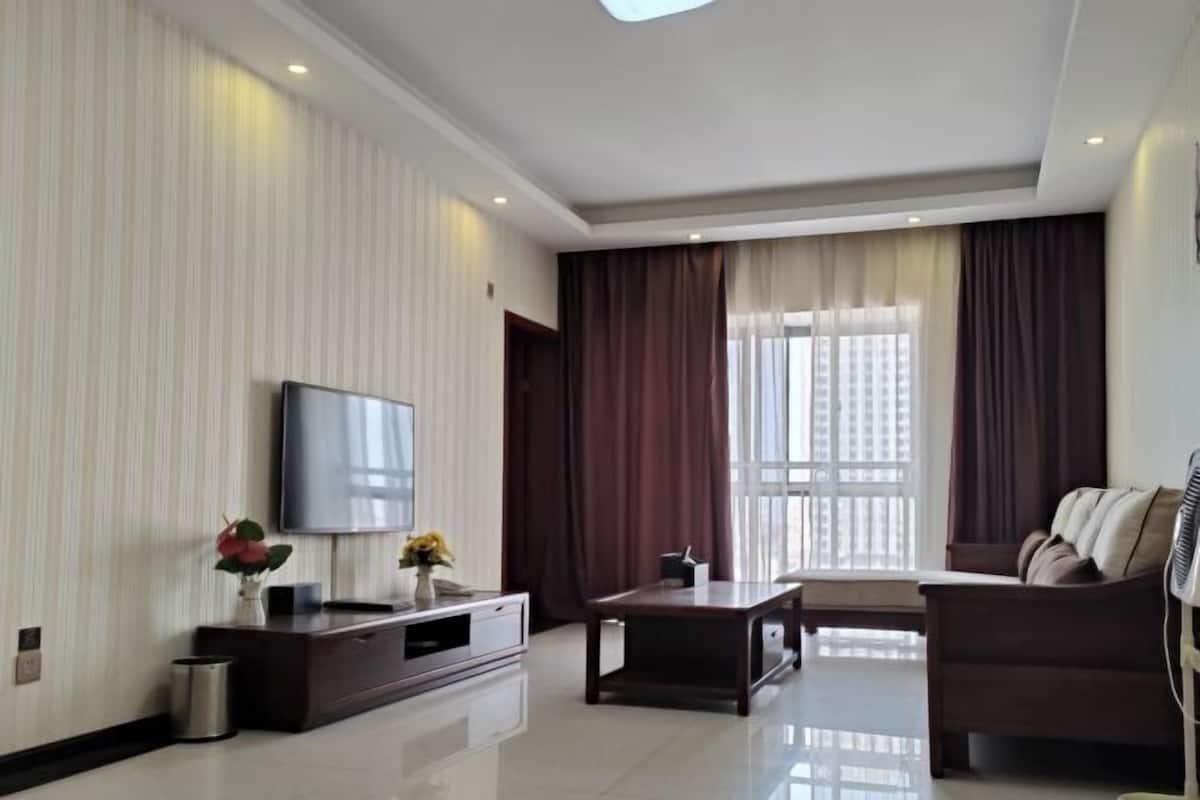 Een moderne woonkamer met een flatscreen televisie, een donkere bank, een salontafel en een groot raam met gordijnen.