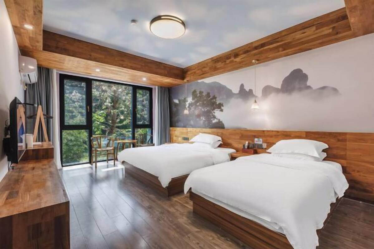 Habitación de hotel con dos camas, techo de madera, un ventanal con vistas a árboles y un mural de montañas.