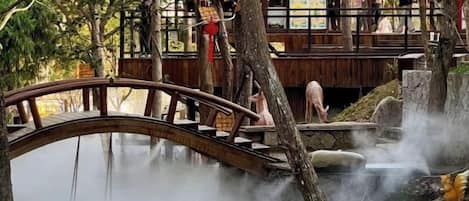 A wooden bridge over a body of water with steam rising from the water, surrounded by trees and a wooden structure in the background.