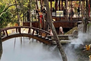 A wooden bridge over a body of water with steam rising from the water, surrounded by trees and a wooden structure in the background. - Puyu Mountain Household (Xianju)