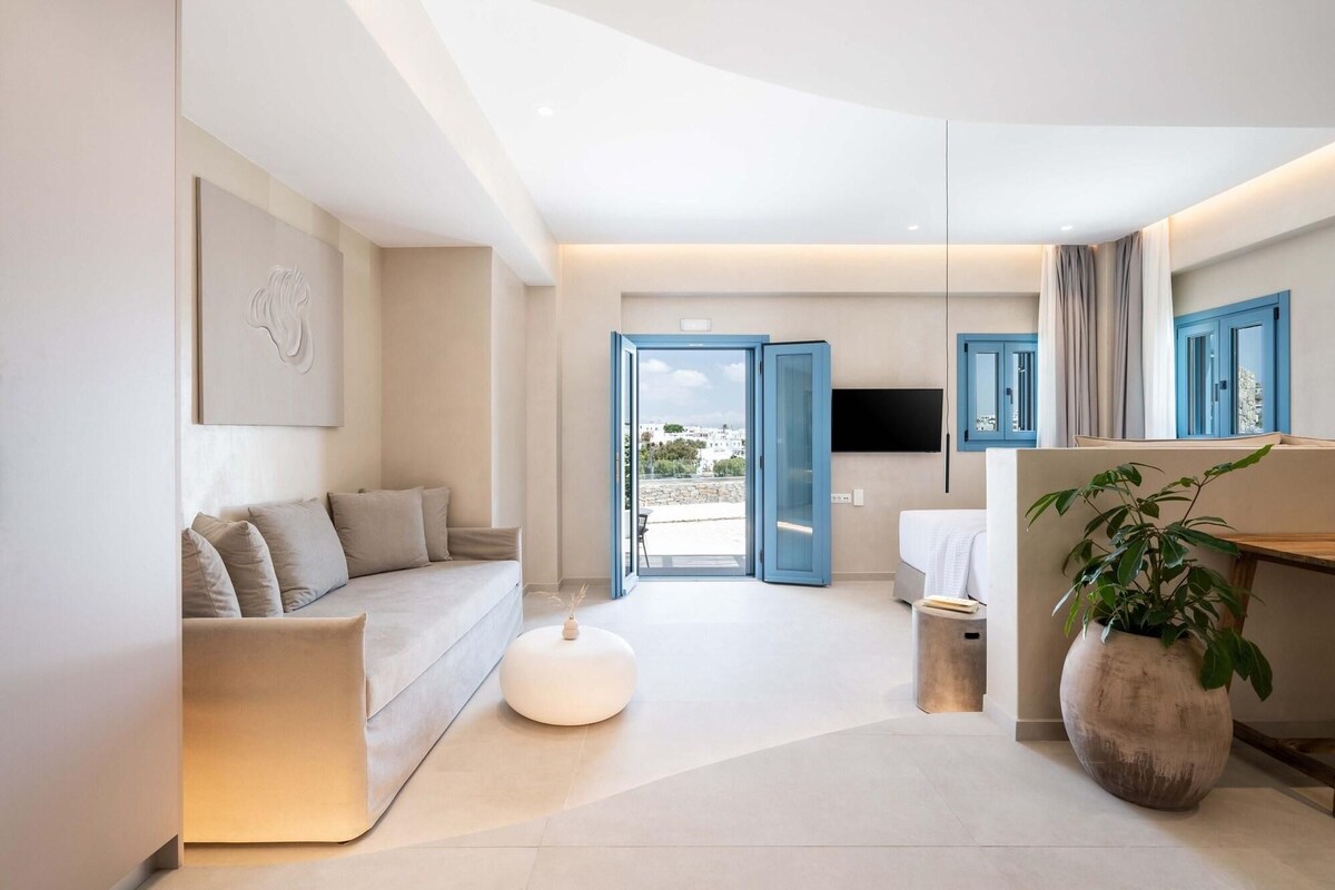 Una habitación de hotel moderna con sofá, un taburete redondo pequeño, una planta grande en maceta, un baño con espejo y vista a la ciudad desde la puerta abierta.