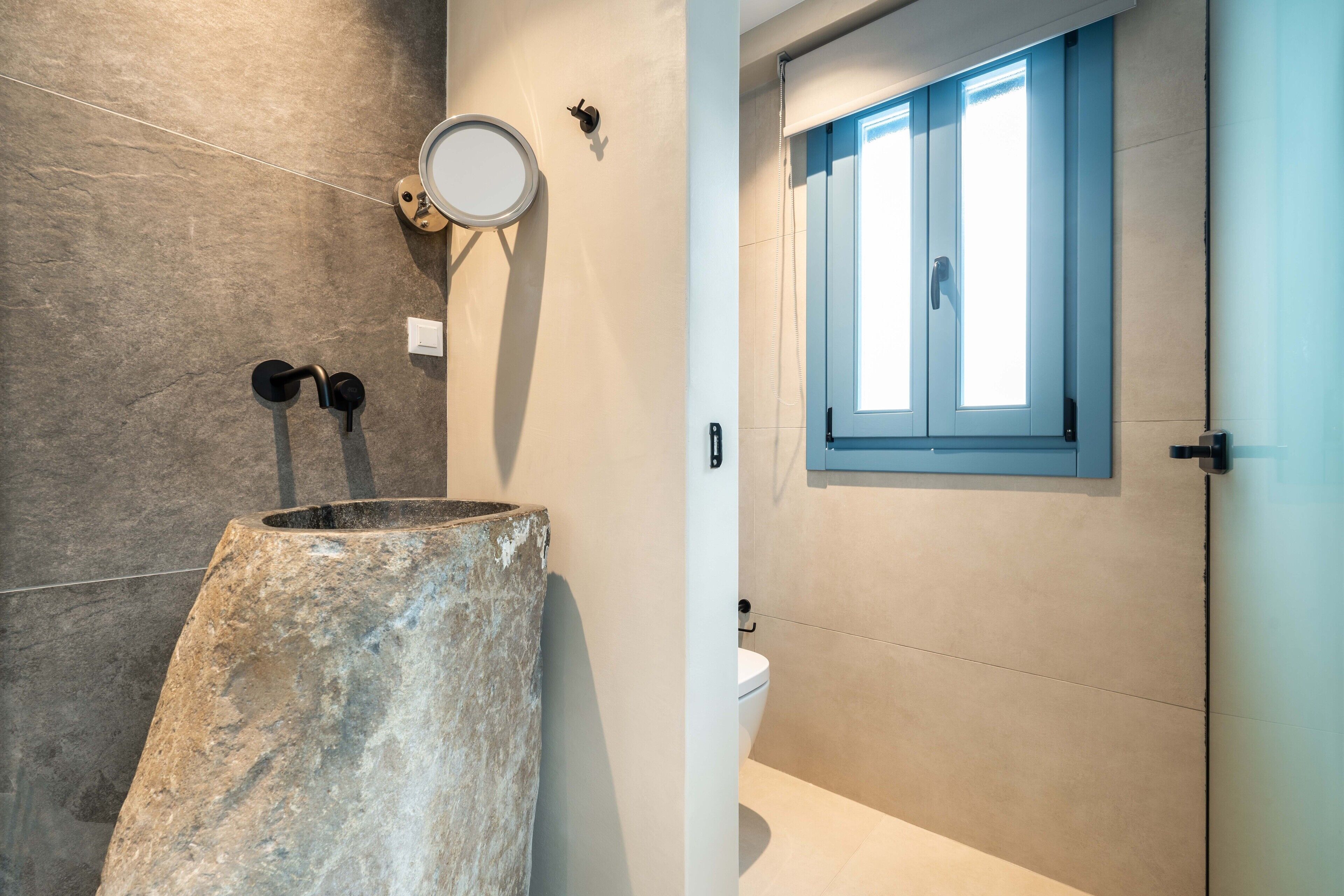 A bathroom with a stone sink, a toilet, and a window with blue shutters.