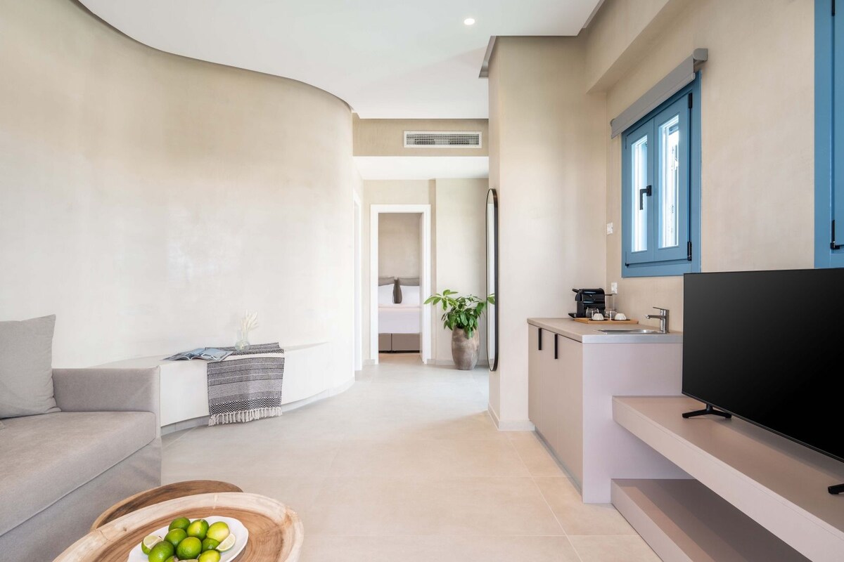 Una sala de estar moderna con un sofá, televisor y una zona de cocina con lavabo y una planta.