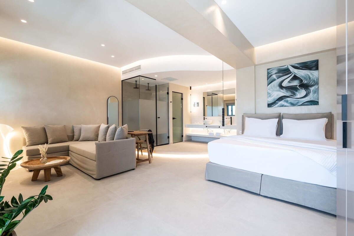 A modern bedroom with a large bed, a sofa, a dining table, and a bathroom with a large mirror.