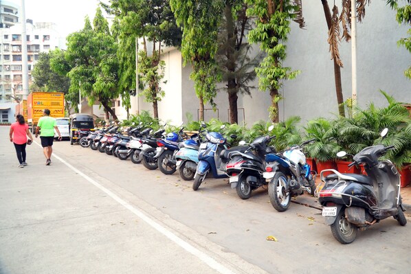 Une rangée de scooters garés le long d’un trottoir, avec des arbres et un immeuble en arrière-plan.