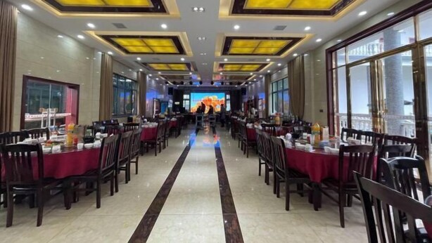 A spacious dining area with tables set for multiple people, large windows on one side, and a central stage with a large screen. - Jiayuan Holiday Hotel (Liuyang)