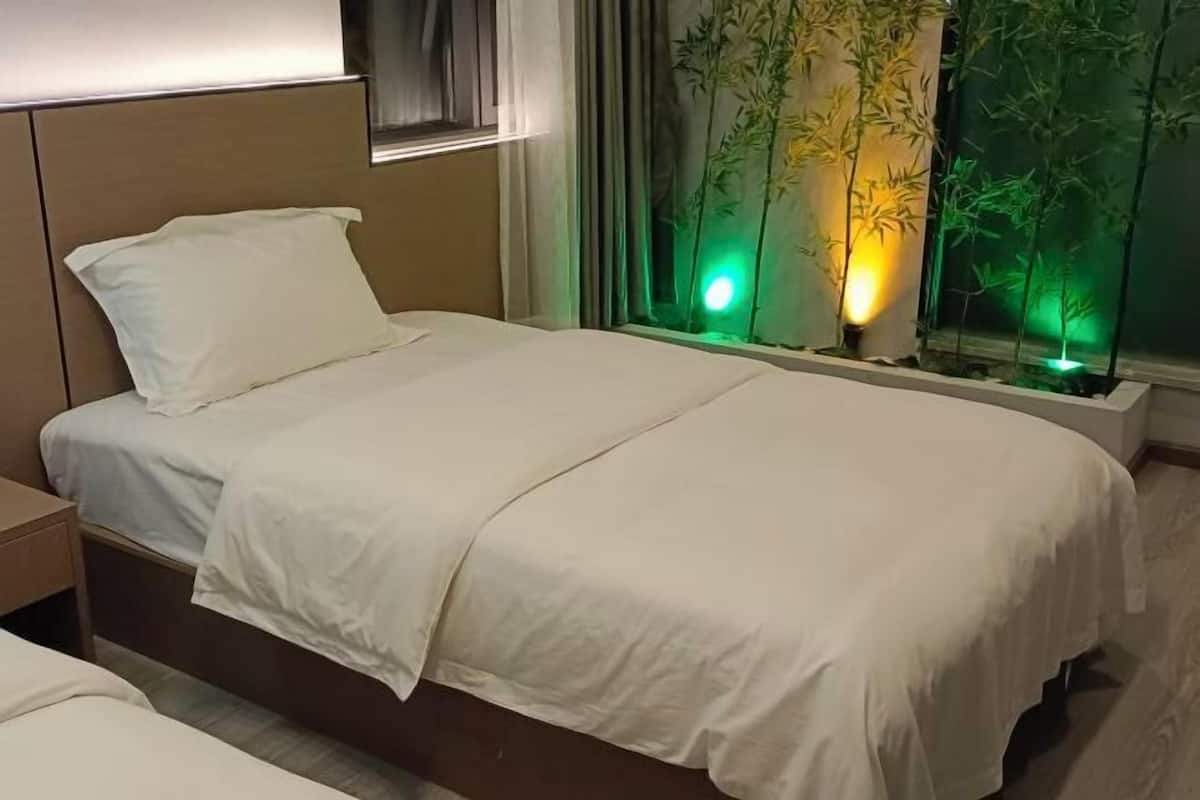 A neatly made bed with white linens in a modern room with a window featuring green lighting and bamboo plants.