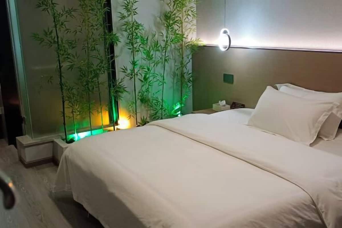 A hotel room with a bed, a nightstand, a wall-mounted lamp, and a bamboo plant.