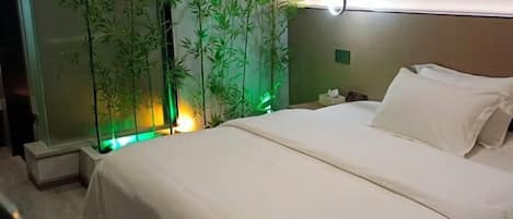 A hotel room with a bed, a nightstand, a wall-mounted lamp, and a bamboo plant.