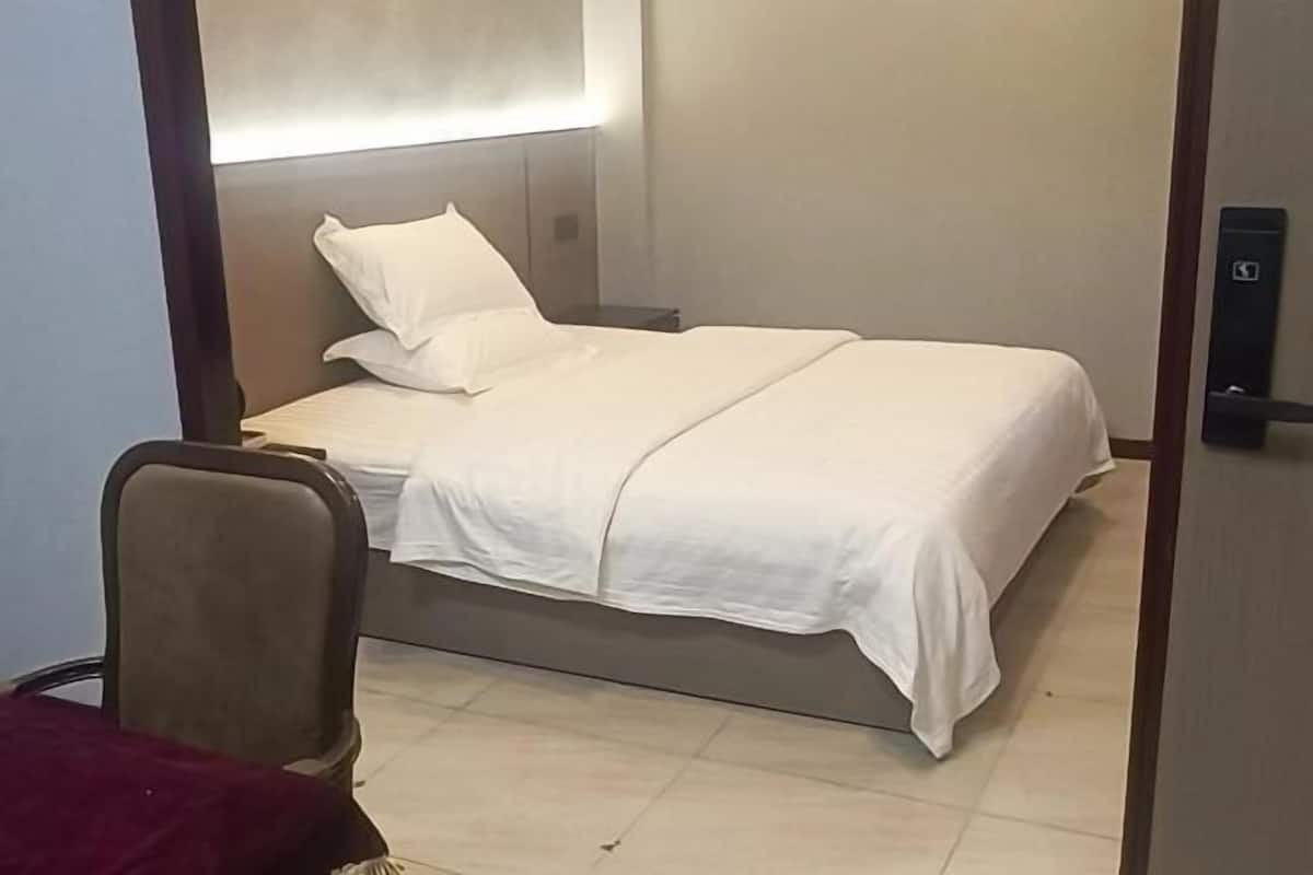 A single bed with white bedding, a nightstand, and a chair in a room with a plain wall and a door.