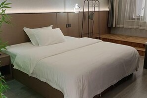Un lit bien fait, avec du linge de lit blanc, une table de chevet, un luminaire fixé au mur et une fenêtre ornée de rideaux légers.