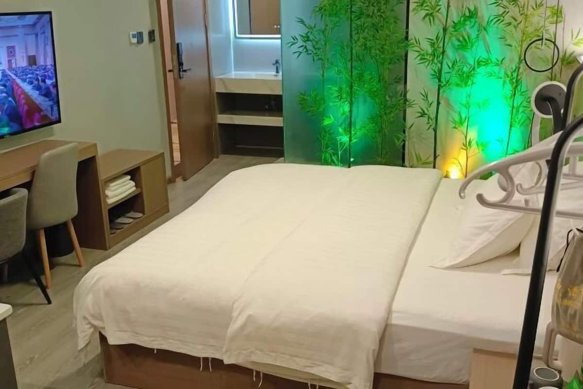 A hotel room with a bed, a desk with a chair, a television, and a bamboo plant.