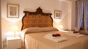 A neatly made bed with a floral headboard, two bedside lamps, and a stack of folded towels with red flowers. - Romantic apartment with canal view (Venezia)