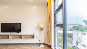 A room with a TV mounted on the wall, a floor lamp, and a window with a view of buildings and water. - Time Walk Guesthouse (Shenzhen)