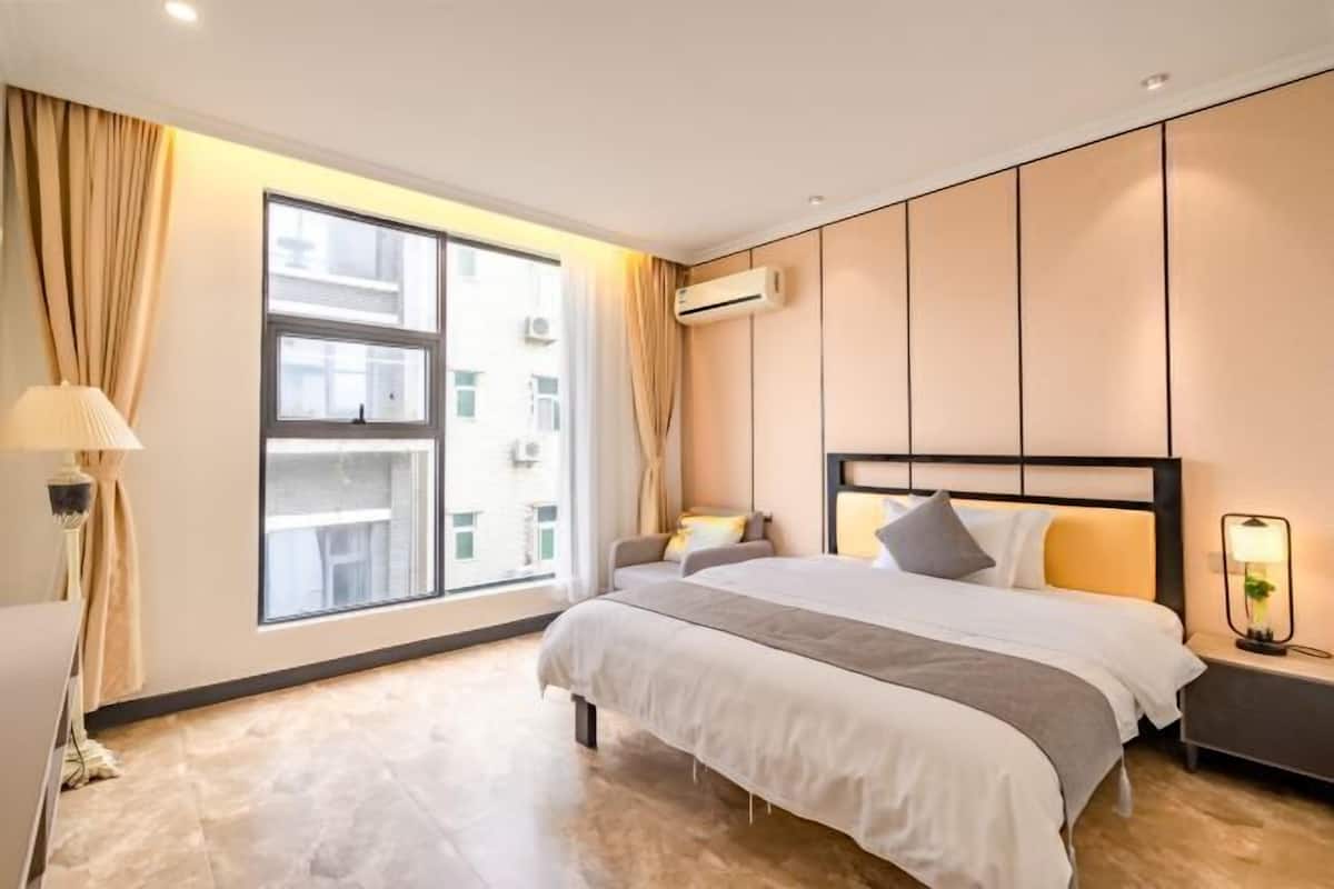A modern hotel room with a large window, a bed with a headboard, a bedside table, and a lamp.