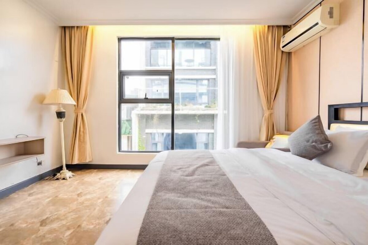 A hotel room with a large bed, a standing lamp, a view of buildings through a window, and an air conditioning unit.