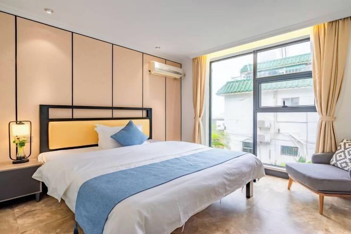 A hotel room with a large bed, a nightstand with a lamp, a window with curtains, and a view of buildings outside.