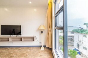 A room with a flat-screen TV mounted on the wall, a floor lamp, and a window with a view of buildings and a body of water. - Time Walk Guesthouse (Shenzhen)