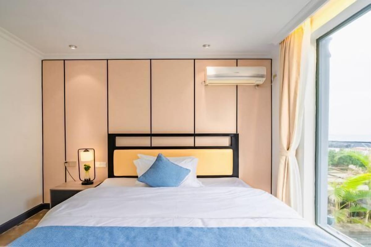 A hotel room with a bed, a nightstand, an air conditioning unit, and a window with curtains.