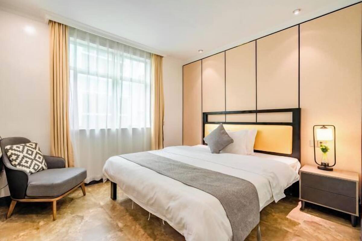 A hotel room with a large bed, a chair, a bedside table with a lamp, and a window with curtains.