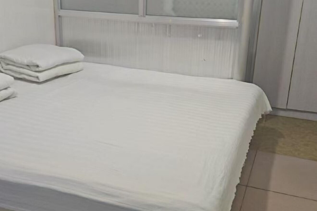 Un lit bien fait, recouvert d’une courtepointe blanche et agrémenté d’oreillers, une fenêtre avec des stores et un sol carrelé.
