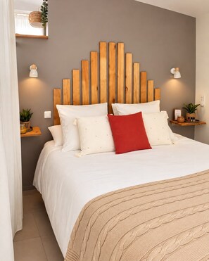 Un lit avec une tête de lit en bois, une literie blanche et un coussin rouge décoratif. On y voit des lampes fixées au mur et une petite plante posée sur une table de chevet.