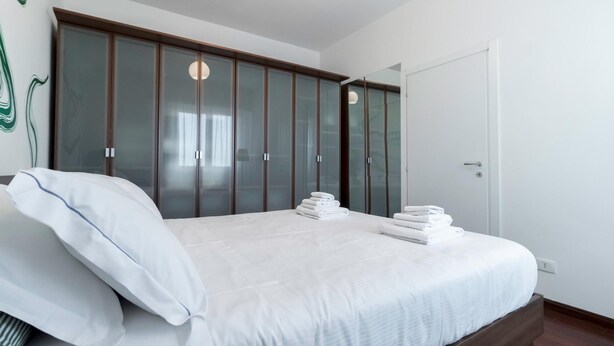 Quarto com cama, roupa de cama branca, guarda-roupa de madeira e espelho.
