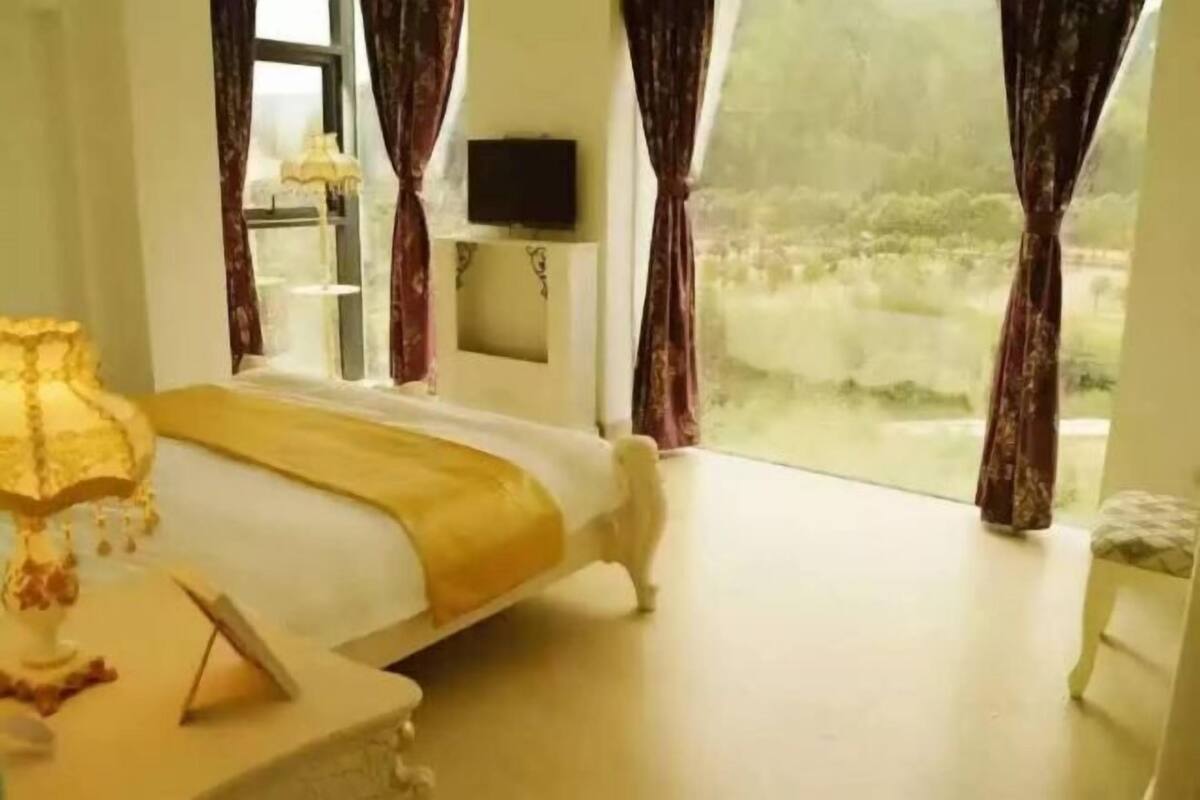 Een slaapkamer met een bed, nachtkastje, lamp, televisie aan de muur en een raam met gordijnen.