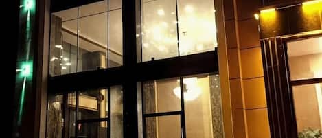 Fachada de edificio con grandes ventanales, un interior visible con un candelabro y una puerta con una persona en la entrada.