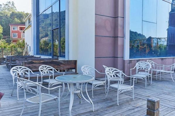 Un área de estar al aire libre con sillas y mesas de metal blanco, un banco de madera y un amplio ventanal que refleja un paisaje montañoso.