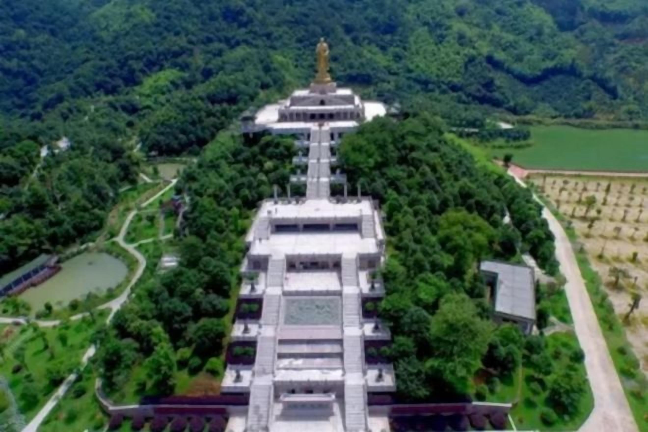 A large, multi-tiered structure with a central statue, surrounded by lush greenery and a body of water.