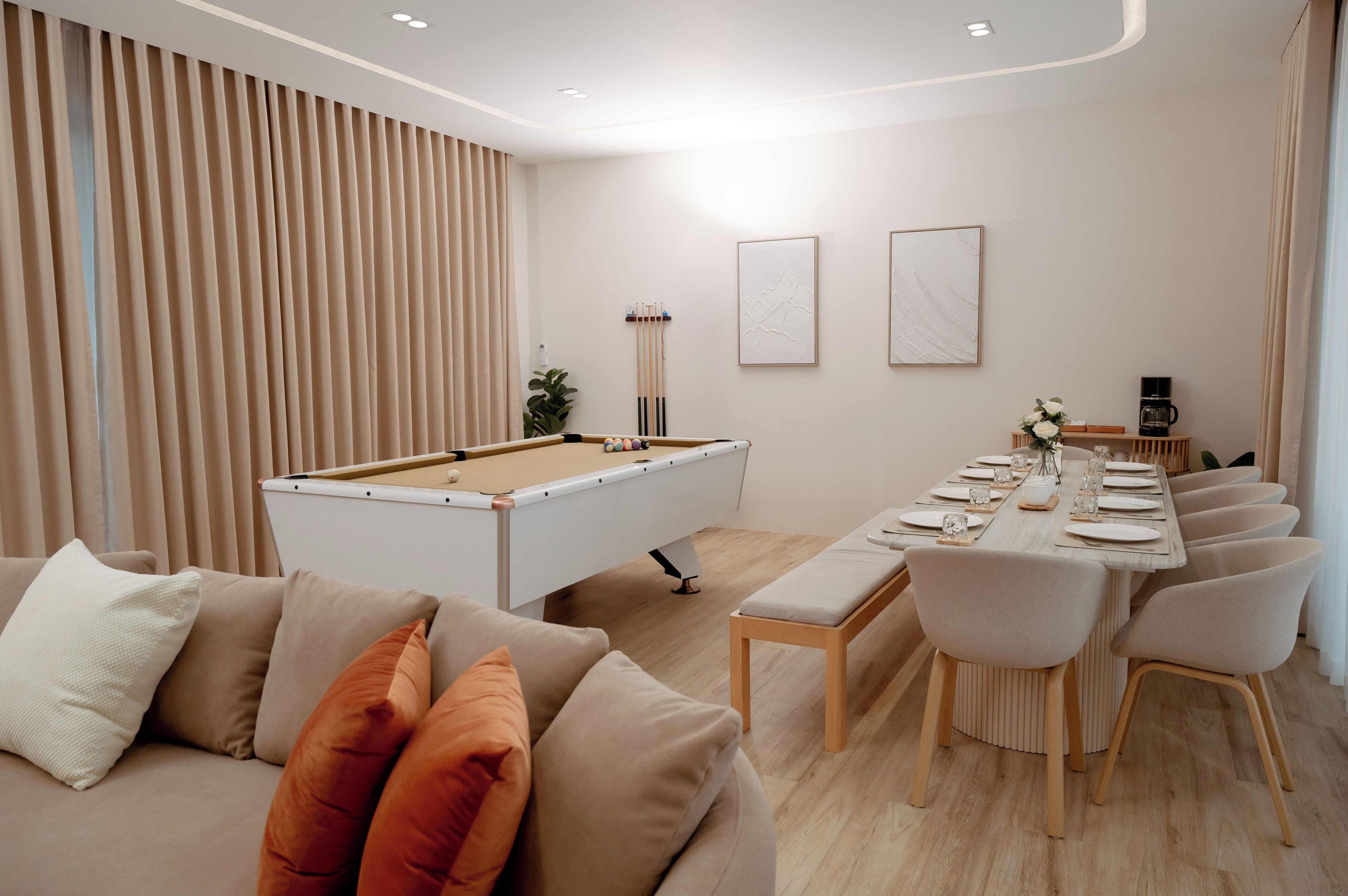 A modern living room with a pool table, a dining area with a long table set for six, and a sofa with cushions.