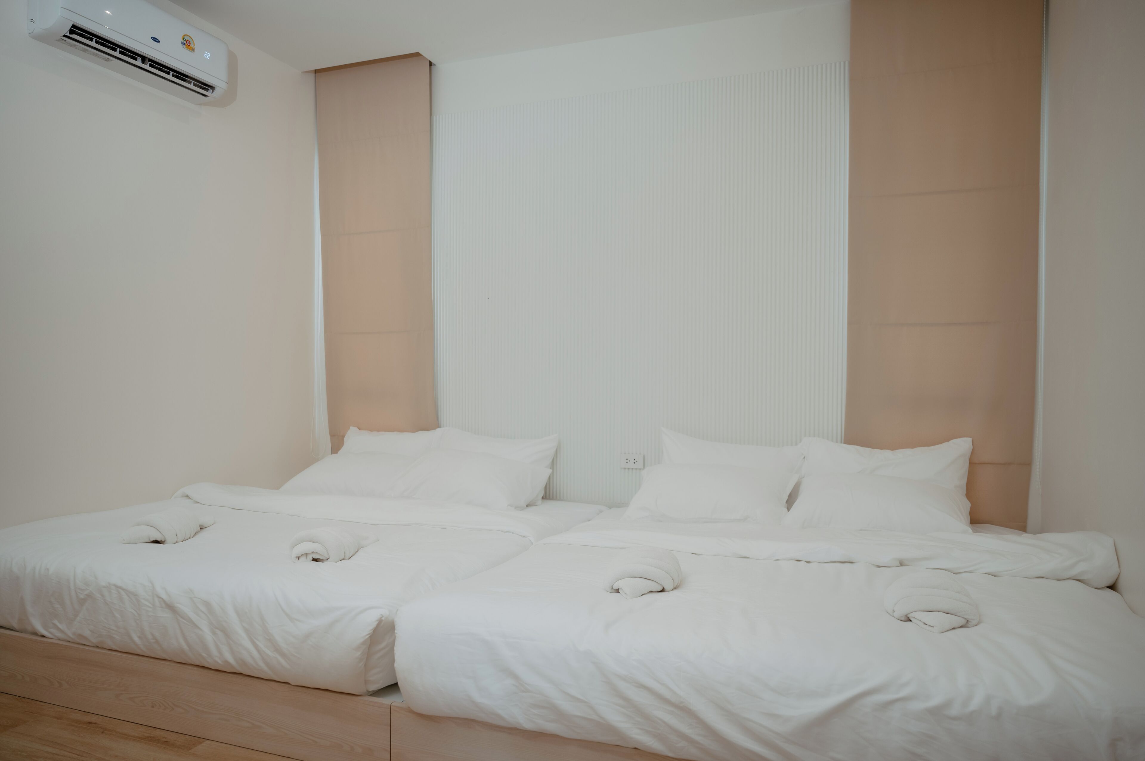 A double bed with white bedding and a headboard, an air conditioning unit above it, and a light-colored wall.