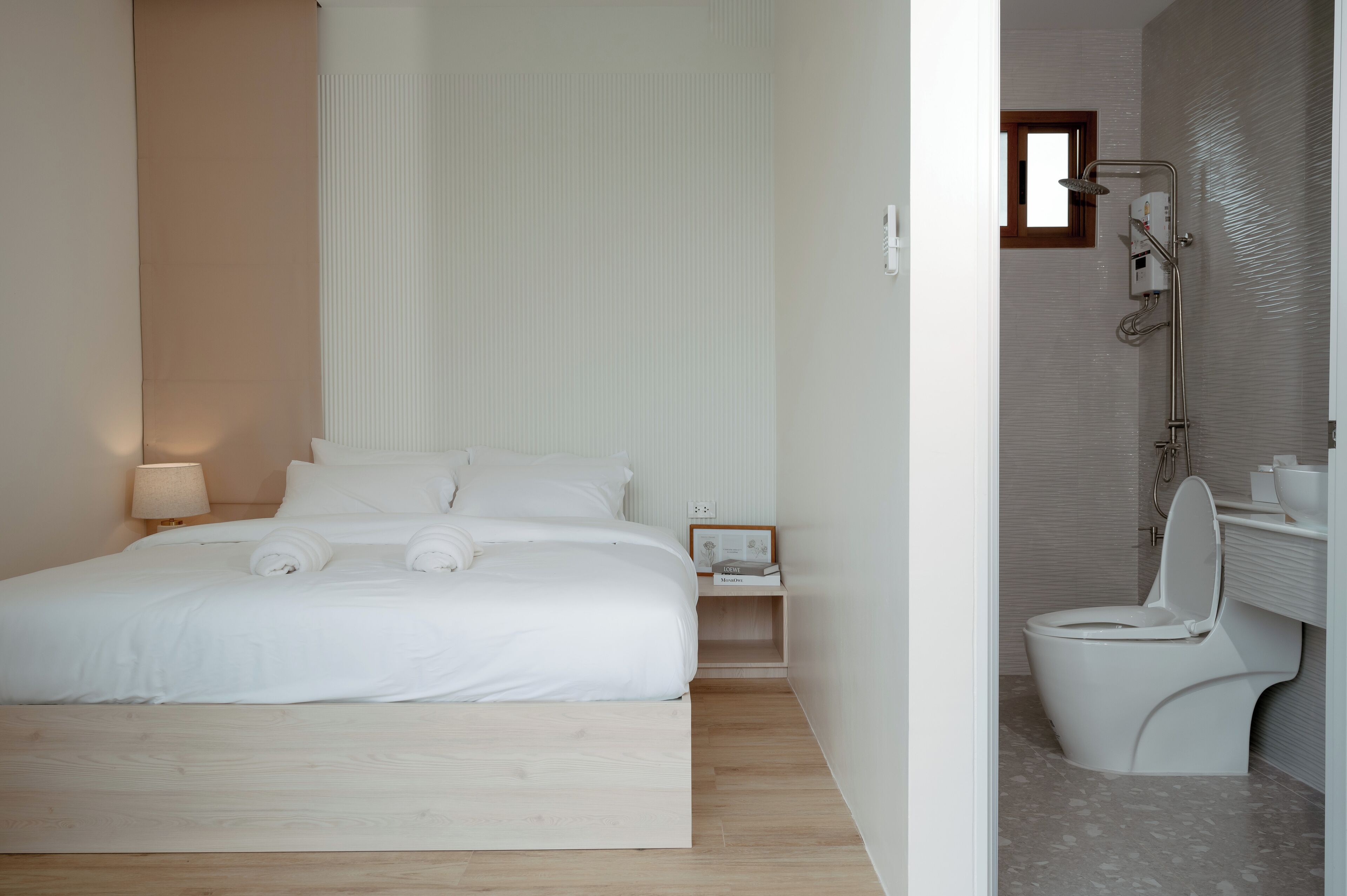 A modern hotel room with a large bed, a bedside table with a lamp, a small bedside table with a book, and a bathroom with a toilet and shower.