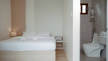 A modern hotel room with a large bed, a bedside table with a lamp, a small bedside table with a book, and a bathroom with a toilet and shower.