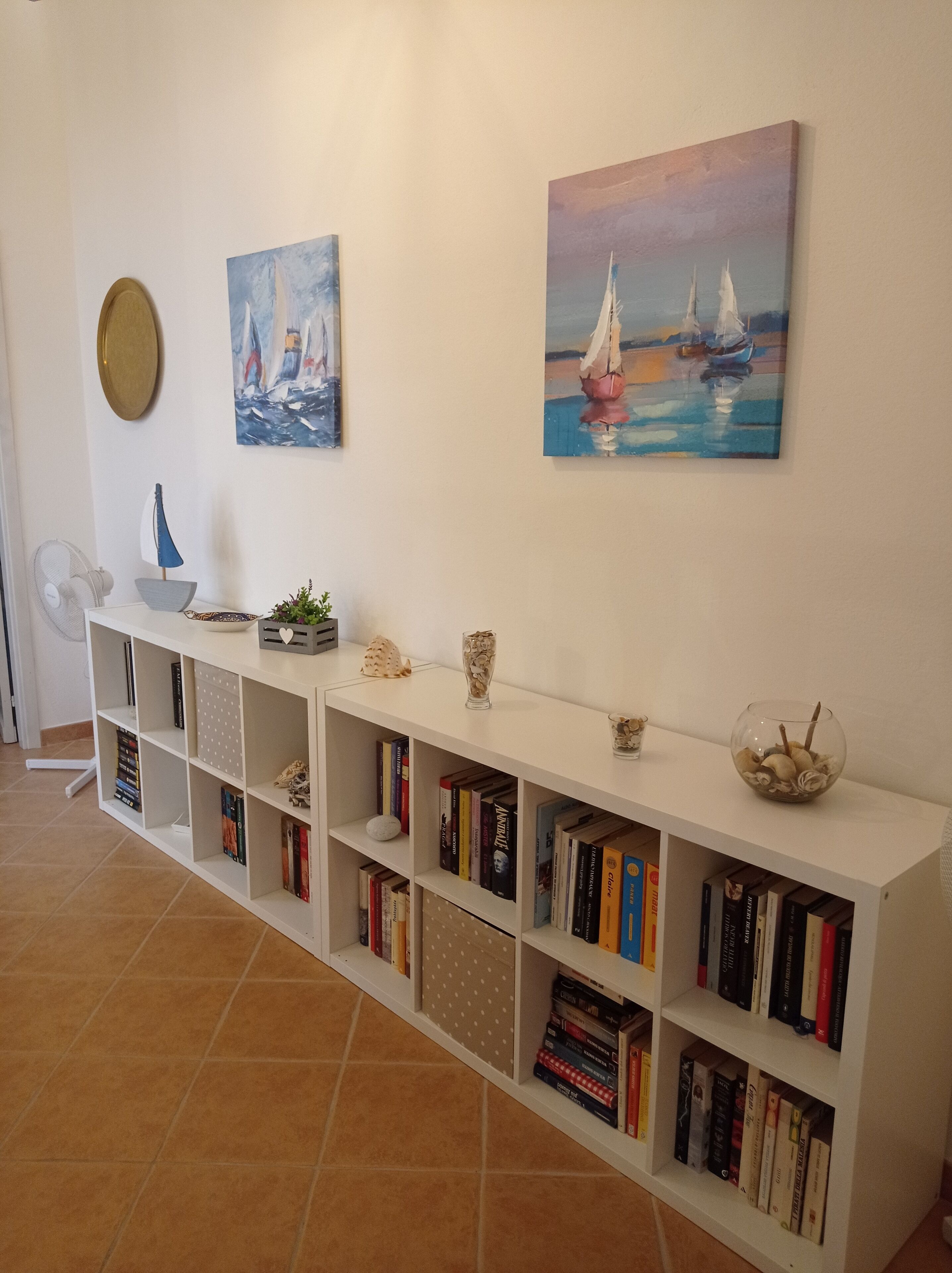 A white bookshelf with books and decorative items, a small fan, and two paintings of sailboats on the wall.